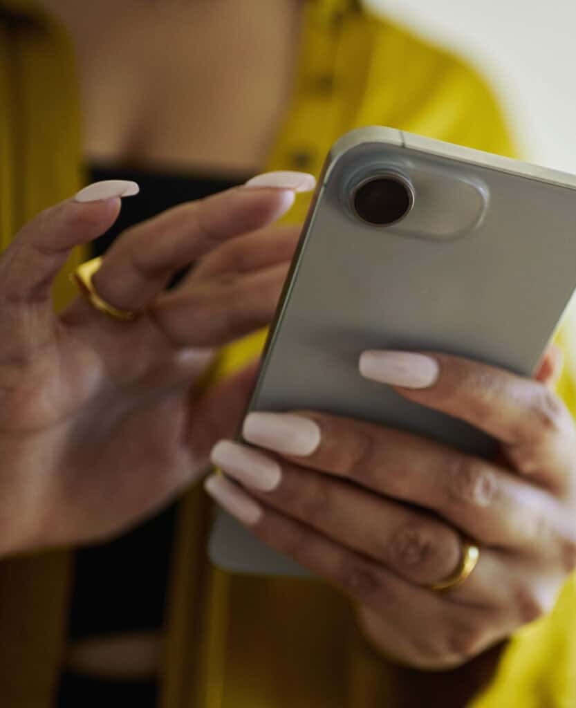 Flexible smartphone camera close-up, person using mobile phone, silver device, modern technology, smartphone photography, hands and nails visible, casual yellow outfit, digital communication, everyday mobile use.