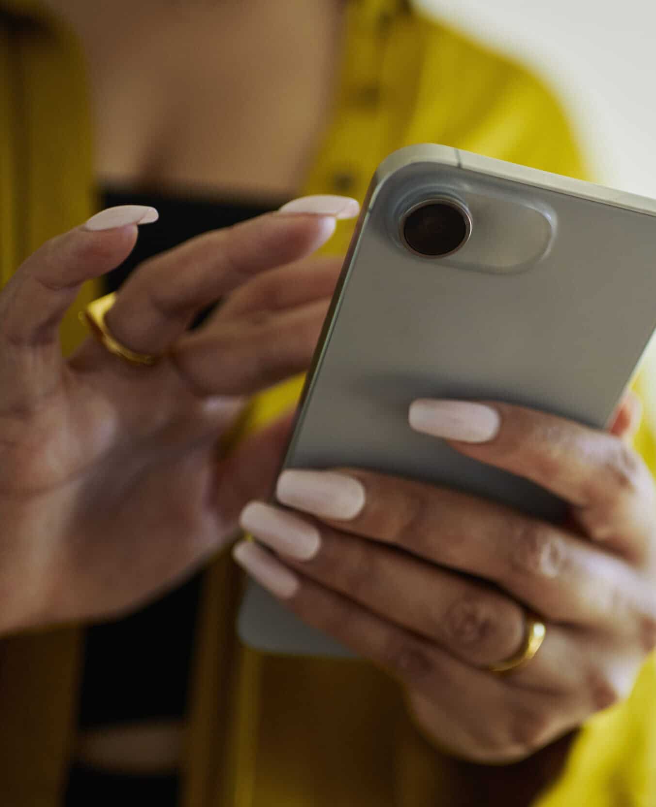 Flexible smartphone camera close-up, person using mobile phone, silver device, modern technology, smartphone photography, hands and nails visible, casual yellow outfit, digital communication, everyday mobile use.