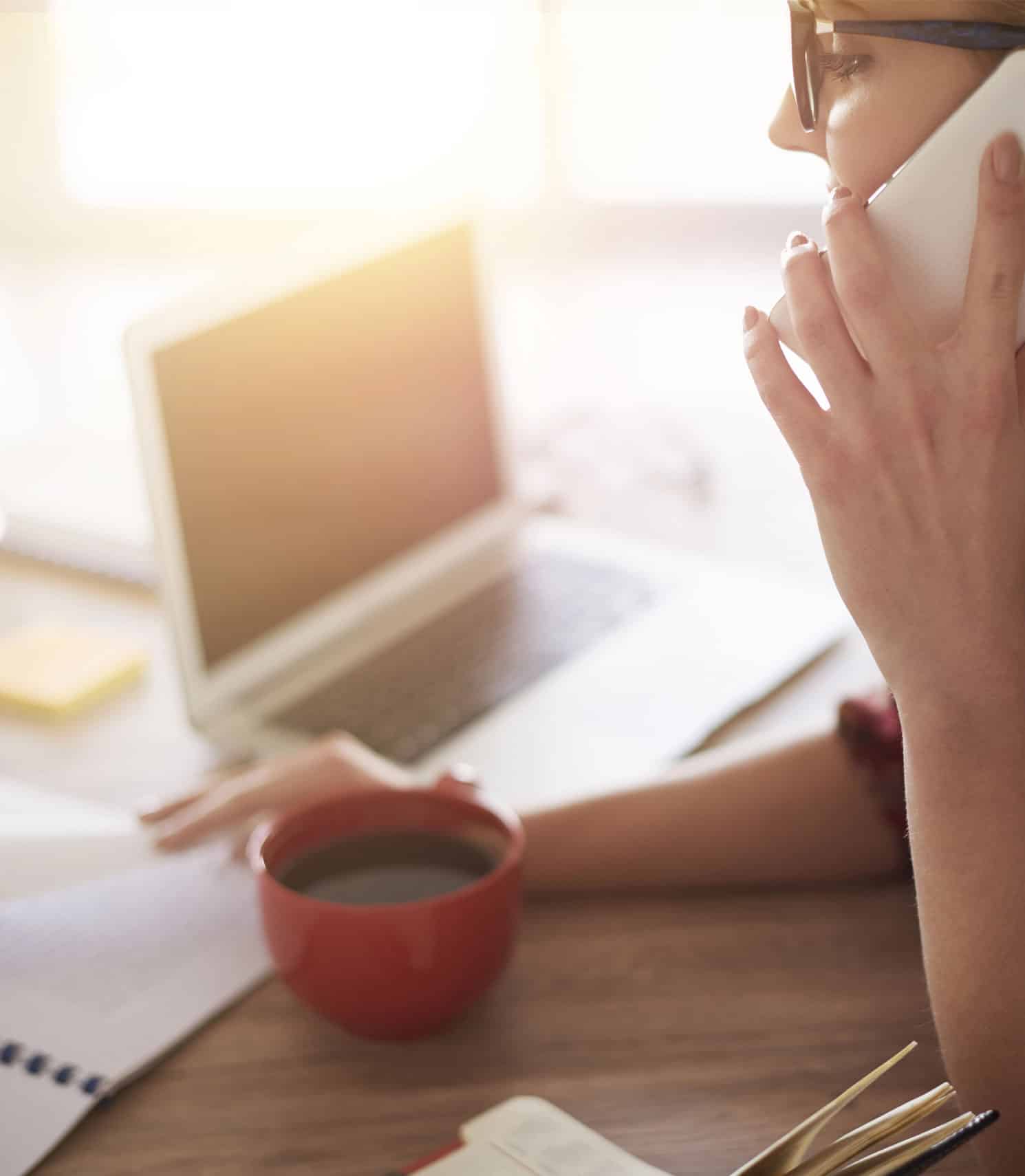 Professional woman using a smartphone and working on a laptop at her desk, representing digital marketing and online business services from Prestige Digital.